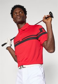 Červený golfový polokošeľ s čiernymi prúžkami cez hruď, s klasickým golierom a textúrovanou tkaninou. V kombinácii s bielymi nohavicami a rukavicou.