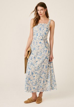 Mujer con un vestido midi blanco sin mangas con estampado floral azul y cinturón, sosteniendo un bolso de flecos color tostado y usando zapatos slip-on color tostado.