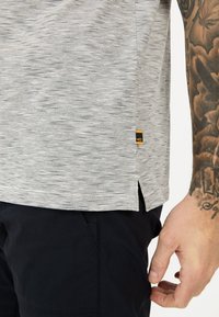 Chemise à manches courtes grise et blanche rayée avec une fente sur le côté et une petite étiquette de marque jaune, faite d'un tissu doux et texturé.
