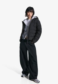 Zwarte pufferjas met een capuchon en gekwalteerde textuur, gelaagd over een lichtgrijze hoodie en gecombineerd met losse zwarte broek en grijze sneakers.