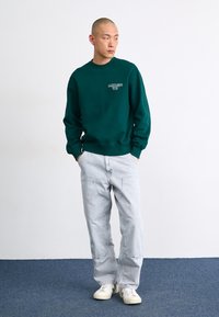 Grøn sweatshirt med rund hals og logo, parret med lyseblå jeans. Modellen bærer hvide sneakers. Enkel design, afslappet pasform.