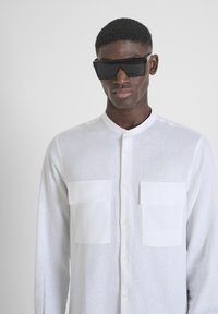 Camicia bianca di lino con maniche lunghe, colletto arrotondato, due tasche frontali e chiusura a bottoni. Il modello indossa occhiali da sole neri oversized.
