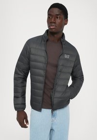 Mann som har på seg en svart quiltet jakke med logo, brun skjorte og lyseblå jeans, står med en hånd i lommen og ser til siden.