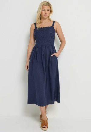 Frau trägt ein marineblaues Midikleid mit floraler Spitzenoberteil und Trägern, Hand in der Tasche, kombiniert mit braunen offenen Keilsandalen.