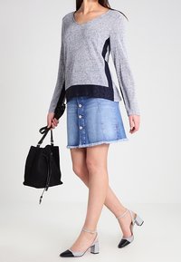 Graues Langarmshirt mit marineblauen Akzenten, kombiniert mit einem hellblauen Jeansrock mit Knöpfen vorne und Fransen am Saum. Schwarze Handtasche und graue Sandaletten mit Absatz.