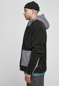 Jachetă din fleece cu un exterior texturat negru, glugă gri și buzunare. Designul include un fermoar frontal și un panou gri contrastant.