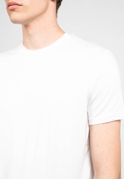 T-shirt bianca in cotone con scollatura rotonda. Maniche corte e una texture liscia. Inquadratura ravvicinata che mette a fuoco l'area del collo e della spalla.
