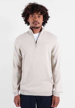 Pull en tricot beige avec une fermeture éclair quart, des poignets côtelés et un détail de logo subtil. Texture douce, coupe régulière, conçu pour le confort.