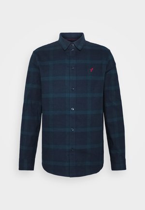 Chemise à manches longues à carreaux bleu foncé et noir avec des boutons et un petit logo rouge brodé sur la poitrine gauche.
