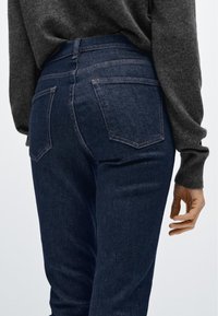 Mörkblå jeans i denim med en figurnära design, som har två bakfickor och diskreta söm-detaljer. Bärs med en grå texturerad tröja.