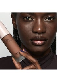 Gros plan d'une femme à la peau foncée tenant une bouteille de fond de teint KIKO Milano Love Fusion près de son visage, montrant une peau lisse et des lèvres brillantes.