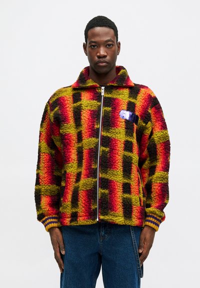 Raudonas, geltonas ir juodas pledinės medvilnės striukė su užtrauktuku priekyje, apykakle ir įsiūtomis rankogaliais. Apykaklėje yra mėlynas logotipas.