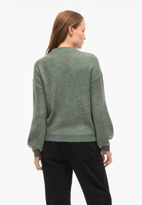 Zielony dziergany sweter o prążkowanej fakturze, z okrągłym dekoltem i oversize'owymi rękawami. W komplecie z czarnymi spodniami, pokazany od tyłu.