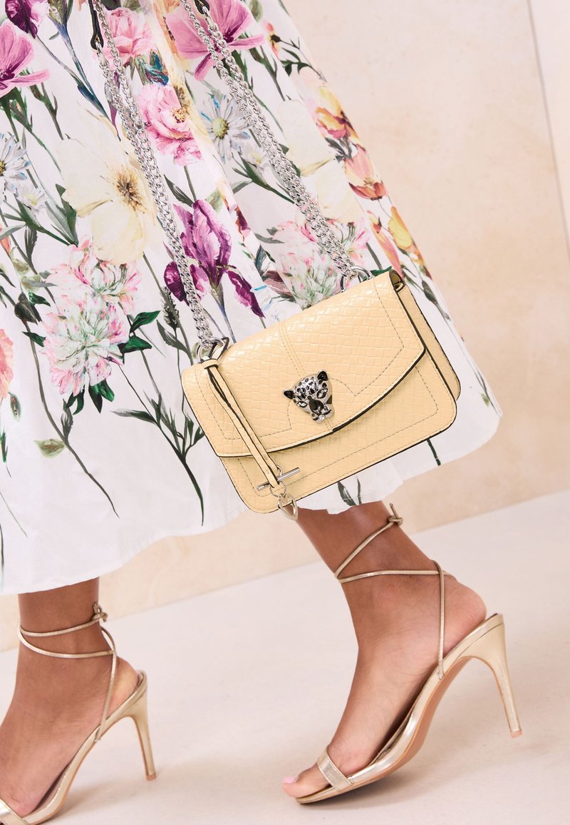 Bolso beige texturizado con cierre de leopardo y cadena plateada, combinado con sandalias doradas de tiras con tacón alto y un vestido blanco con estampado floral.