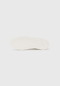 Suela de zapatilla blanca con patrones texturizados, que presenta ranuras horizontales y verticales para mayor agarre. Superficie lisa y diseño de punta redondeada.
