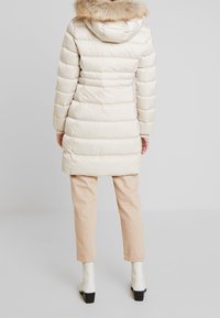 Puffer coat in cream with a fur-trimmed hood, featuring horizontal quilted design. Paired with light beige pants and white ankle boots.