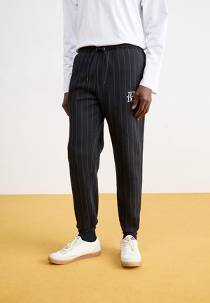 Homme portant un jogging noir à fines rayures avec le logo « JUST DO IT » et des baskets blanches sur un sol beige contre un mur blanc.