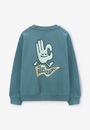 Sweatshirt teal présentant un large graphisme au dos avec un symbole de main et le texte "LES RÈGLES NE S'APPLIQUENT PAS" en jaune et noir.