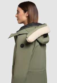 Olivovo zelený park s čiernou podšívkou, plyšovým krémovým golierom a veľkými prednými vreckami. Obsahuje uzávery na gombíky a kapucňu.