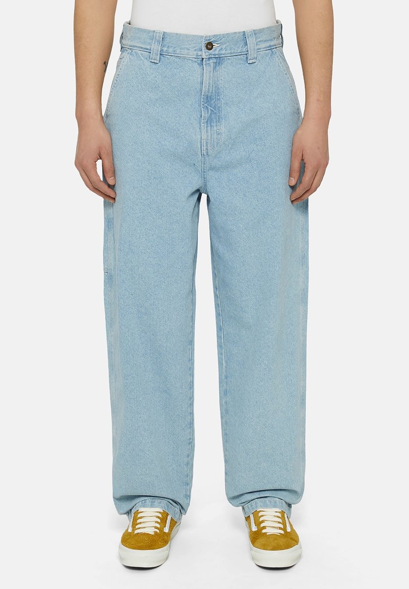 Personne portant un jean bleu clair ample et des baskets en daim jaune avec des lacets blancs, debout devant un fond uni.