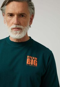 Camiseta de manga larga verde azulado oscuro, hecha de algodón, con una impresión naranja que dice "DREAM BIG" en el lado izquierdo del pecho. Textura suave, cuello redondo.