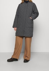 Cappotto grigio trapuntato con collo alto, zip frontale e tasche laterali, abbinato a pantaloni a gamba larga color cammello e scarpe scure.