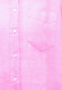 Prim-plan al părții frontale a unei cămăși roz din in, cu nasturi albi și un singur buzunar la piept pe partea dreaptă.