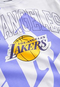Hvid T-shirt med "LOS ANGELES" i lilla fede bogstaver og "LAKERS" logoet med en gul basketball på en lys lilla baggrund.