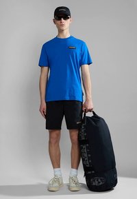 Kék rövid ujjú póló logóval, fekete rövidnadrág, fehér zokni, napszemüveg, sapka, és egy nagy fekete sporttáskát visel, amin grafika látható.