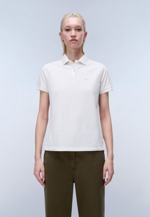 Young person with short blonde hair wearing a white short-sleeve polo shirt and olive green pants, standing against a plain light background.