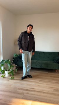 Casaco preto com bolsos, t-shirt gráfica, jeans azul-claro e sapatos pretos, de pé sobre um chão de madeira perto de um sofá verde e uma planta.