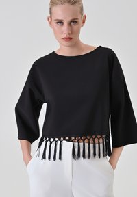 Top nero corto con maniche larghe, caratterizzato da un dettaglio a frangia annodata lungo l'orlo. Tessuto liscio e lucido con finitura opaca.