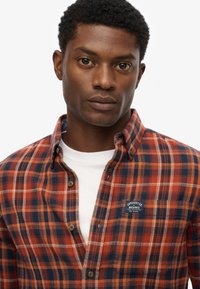 Plaid flannel shirt in orange, navy, and white. Features a classic collar, button closure, and logo patch on the chest. Textured fabric.