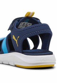 Marinblå sandaler med justerbara remmar, texturerad ovandel, vit mellansula och gul yttersula. Logotypen är synlig på sidan.