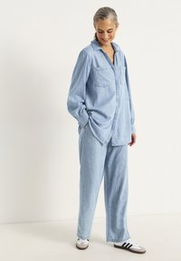 Ljusblå denimskjorta med två bröstfickor, avslappnad passform, långa ärmar, kombinerad med matchande ljusblå denimbyxor och vita sneakers.