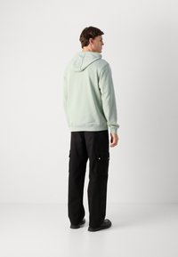 Hombre de pie de lado, vestido con una sudadera verde clara y pantalones cargo negros, con zapatos negros, frente a un fondo liso claro.