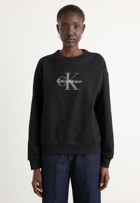 Čierna mikina s okrúhlym výstrihom vyrobená z bavlnenej zmesi, so stredovým bielym a sivým logom "Calvin Klein" a rebrovanými manžetami a spodným lemom.
