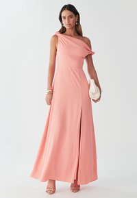 Vienas plecs, korāļrozā maksi vakarkleita ar volānu detaļām, sānu šķēlumu un elegantām līnijām. Aksesuāri ietver teksturētu krēmkrāsas rokassomiņu un siksnu sandales.