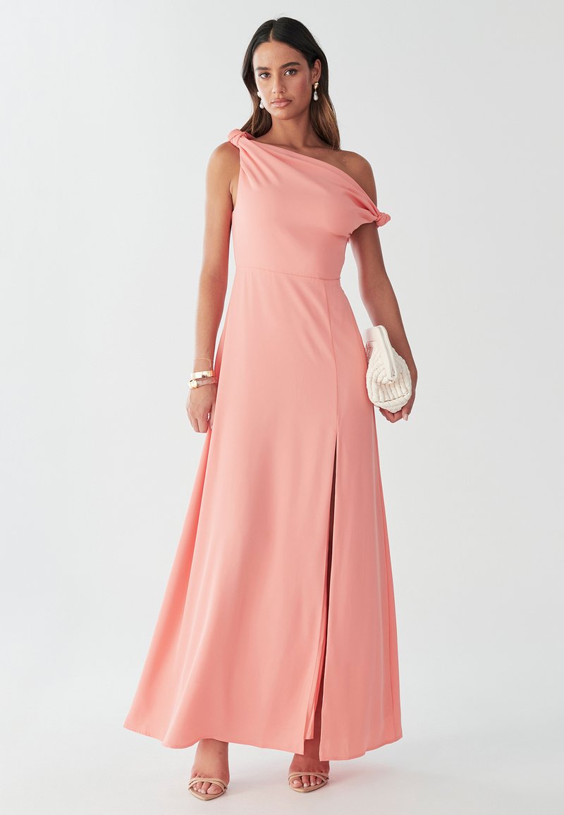 Vienas plecs, korāļrozā maksi vakarkleita ar volānu detaļām, sānu šķēlumu un elegantām līnijām. Aksesuāri ietver teksturētu krēmkrāsas rokassomiņu un siksnu sandales.