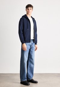 Hanorac navy cu fermoar și șnururi, purtat cu un tricou alb, blugi drepți bleu deschis și pantofi negri, pe un fundal minimalist.