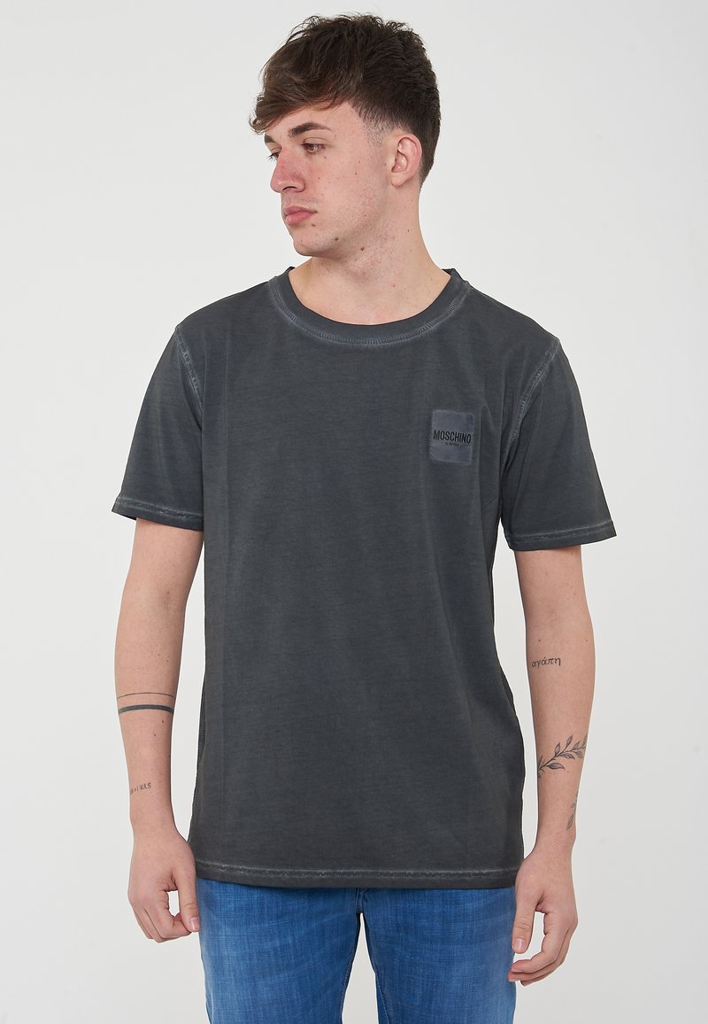 T-shirt in cotone grigio scuro con collo a giro, maniche corte e una toppa testurizzata con il marchio "Moschino" sul petto.