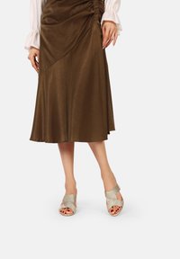 Gonna midi in satin marrone con design diagonale e dettaglio arricciato sul lato. Sandali incrociati metallici con cinturini testurizzati e suola leggera.