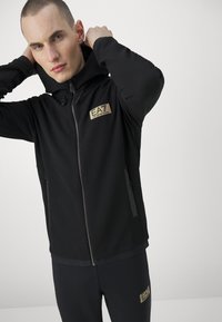 Felpa nero con zip in tessuto texturizzato, cappuccio integrato e due tasche laterali. Presenta un logo dorato sul petto e polsini lisci.