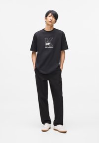 Čierne bavlnené tričko s veľkým bielym logom "K" a grafickým dizajnom, skombinované s voľnými čiernymi nohavicami a bielymi teniskami na neutrálnom pozadí.