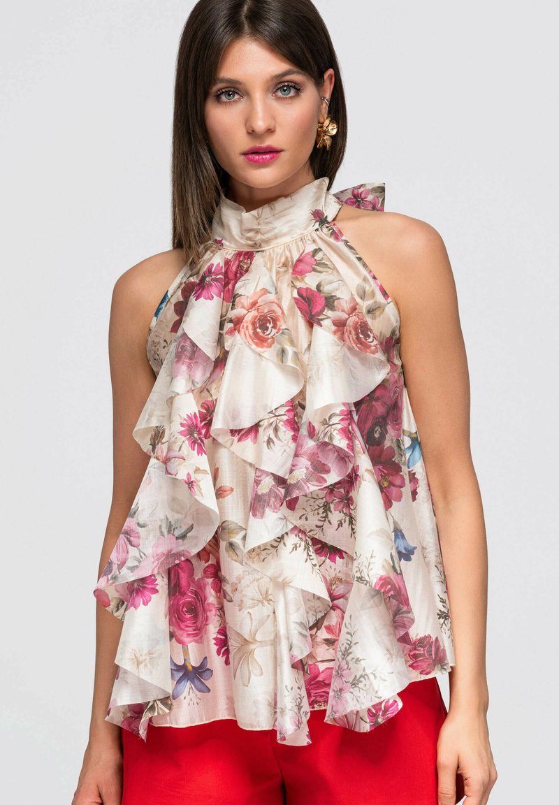 Jeune femme portant un top sans manches à volants fleuri avec des fleurs roses, rouges et beiges, associé à un pantalon rouge vif.