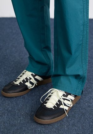 Person iført turkis bukser og sorte sneakers med hvide striber står på et mørkt tæppebelagt gulv.