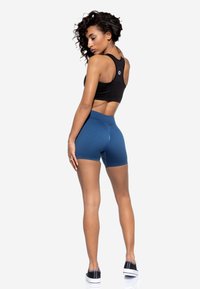 Reggiseno sportivo nero con design racerback abbinato a pantaloni shorts blu a vita alta. Modello che indossa sneakers nere, mostrando il tessuto liscio e la vestibilità.