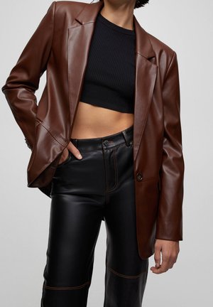 Persona con blazer de cuero marrón, top corto de canalé negro y pantalones de cuero negros con costuras naranjas, de pie frente a un fondo gris.