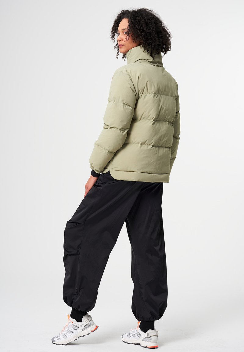 Olivgrüne Daunenjacke mit hohem Kragen, gestepptem Design und schwarzen elastischen Bündchen; kombiniert mit lockeren schwarzen Hosen und weißen Sneakern.