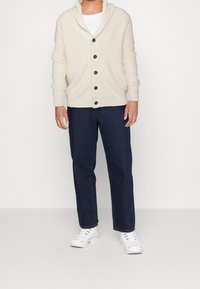 Krémový pletený kardigan se šálovým límcem a hnědými knoflíky, kombinovaný s tmavě modrými džínami s rovnými nohavicemi a bílými teniskami.